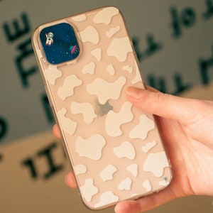 以下が含まれることがあります： 白い牛柄の透明な携帯電話ケース。ケースにはカメラレンズ用の切り抜きと、レンズに小さな宇宙飛行士のステッカーが付いています。