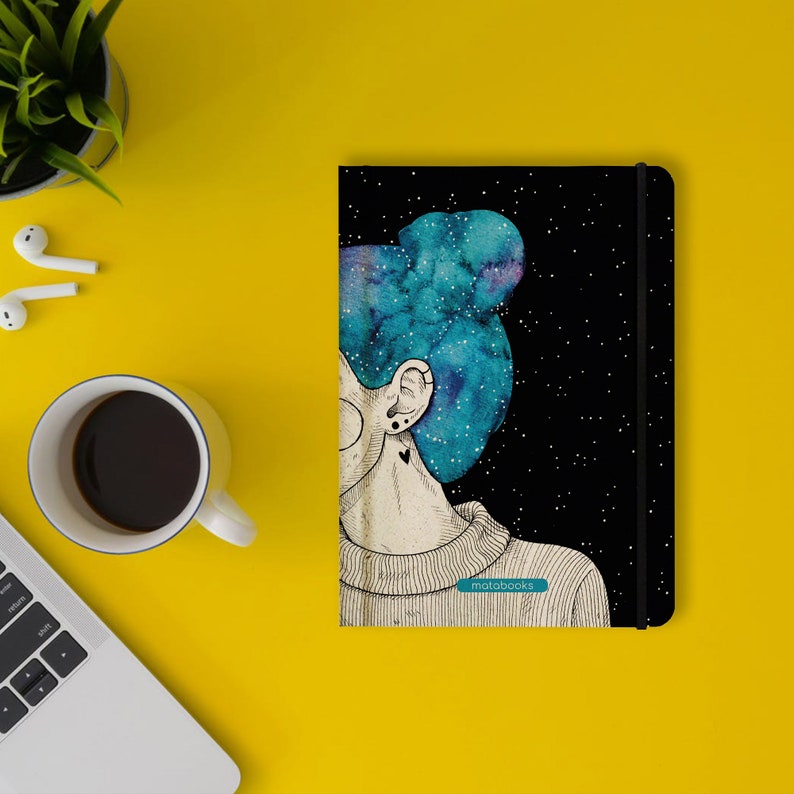 May include: Black notebook with an illustration of a person with blue galaxy-themed hair and a black starry background. The notebook has a black elastic band and the text "motabooks" on the bottom.