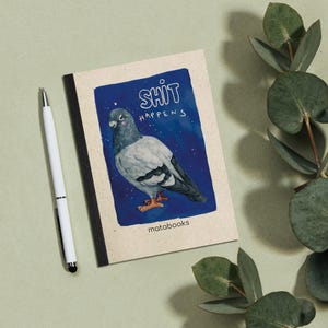 May include: A notebook featuring a blue illustration of a pigeon with the text "SHIT HAPPENS". A white pen lies beside the notebook, which has a black spine. The brand name "matabooks" is printed at the bottom of the illustration.