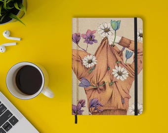 May include: A rectangular notebook with a tan cover featuring a detailed illustration of a person wearing a brown sweater adorned with white daisies and purple tulips. The notebook has a black elastic closure and the word matabooks printed on the front.