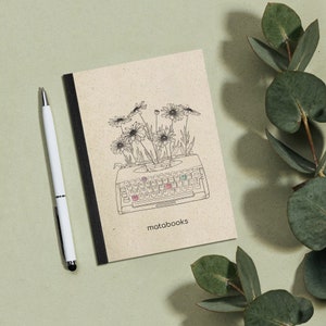 Può includere: Un quaderno marrone con una rilegatura nera e un disegno a linea di una macchina da scrivere con fiori che crescono da essa. Il testo "matabooks" è stampato sulla copertina del quaderno.
