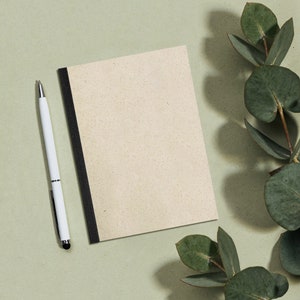 May include: A blank, cream-coloured notebook with a black spine and a white pen lying next to it. The notebook and pen are on a light green surface with green leaves in the background.