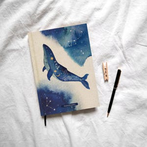 Könnte beinhalten: Ein Notizbuch mit cremefarbenem Einband, das eine blaue Aquarell-Illustration eines Wals mit goldenen Sternen, Monden und Sternbildern zeigt. Das Wort matabooks ist in kleinen Buchstaben unten aufgedruckt. Ein schwarzer Stift und eine Holzklammer liegen daneben.
