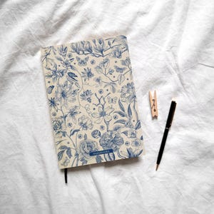 Könnte beinhalten: Ein rechteckiges Notizbuch mit cremefarbenem Einband und einem detaillierten blauen botanischen Druck aus Blumen, Blättern und Schmetterlingen. Das Wort matabooks ist unten in Blau aufgedruckt. Ein schwarzer Kugelschreiber und eine Holzklammer liegen daneben.