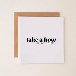 以下が含まれることがあります： 「take a bow - you were amazing」と黒い文字で書かれた白いグリーティングカード。茶色の封筒に入っており、ニュートラルな背景に配置されています。シンプルでエレガントなデザインです。
