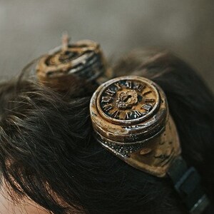 May include: A man wearing steampunk goggles with clock faces on the lenses. The goggles are brown and gold with a black strap.