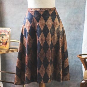 May include: A mannequin displays a brown and navy diamond-patterned skirt. The skirt is knee-length and has a slight A-line shape. The background includes a vintage-style coat rack, a small table, and lace curtains.