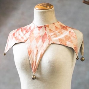 May include: A cream-colored mannequin torso with a jester collar. The collar features a diamond pattern in shades of peach and red, with small gold-colored bells at the points. The mannequin is on a wooden stand.