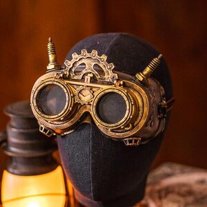 May include: Gold-toned brass goggles featuring a prominent gear mechanism on the bridge, two circular lenses, and small vertical spikes on the sides. The goggles are displayed on a dark fabric-covered head form, showcasing intricate metal detailing.