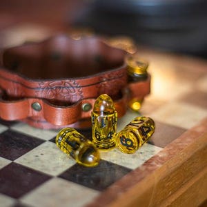 May include: A brown leather bracelet with decorative stitching and brass rivets sits on a chessboard. Three yellow, bullet-shaped objects with black numbers are next to the bracelet. The objects are on a wooden surface.