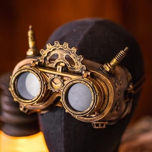 May include: Brass-colored goggles featuring two circular lenses, a central gear mechanism, and two small vertical spikes on the top frame. The goggles are mounted on a dark, featureless head-shaped display form with a metallic, weathered finish.