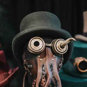 May include: A steampunk-style mask with a top hat, goggles, and an octopus-like design. The mask is made of brown leather and has a dark green top hat. The goggles are made of brass and have round lenses. The octopus tentacles are made of brown leather and have a textured surface.