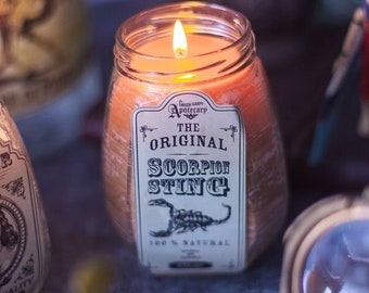 May include: A lit candle in a glass jar with a label that reads "The Original Scorpion Sting 100% Natural". The candle is on a dark gray surface with other objects in the background.
