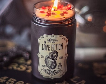 May include: A black candle jar with a label that reads "Green Happy Apothecary Love Potion Est. 1697" with a graphic of a heart. The candle is lit and has red flower petals on top. The candle is sitting on a black surface with gold lettering that reads "4567890 XYZ LM".