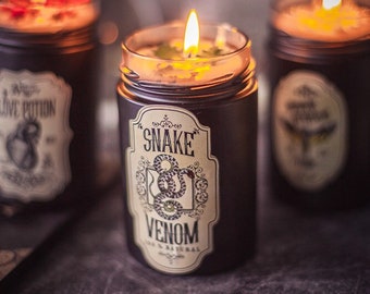 May include: Three black glass candle jars with labels. The center jar has a label that reads "Snake Venom" with a snake illustration. The jar on the left has a label that reads "Love Potion" with a heart illustration. The jar on the right has a label with an owl illustration. All three jars have a lit candle inside.