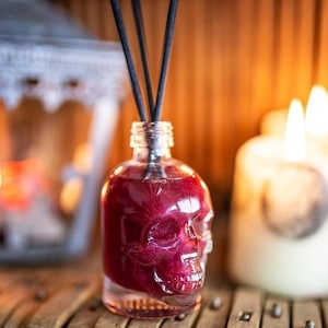 Può includere: Una bottiglia di vetro a forma di teschio riempita di un liquido rosso scuro e bastoncini diffusori neri. La bottiglia è appoggiata su una superficie di legno.