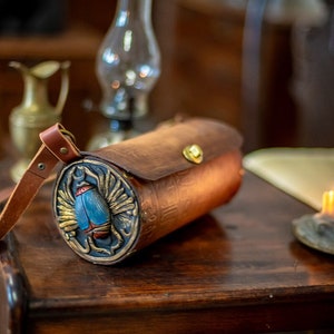 May include: A brown leather cylindrical bag with a strap. The bag has a circular front panel with a gold-colored scarab beetle design with blue wings and a red body.