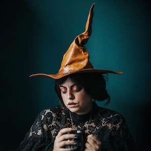 Peut inclure: Une femme portant un chapeau de sorcière pointu brun et une robe en dentelle noire tient une tasse noire avec le texte "Witches Brew" en blanc.