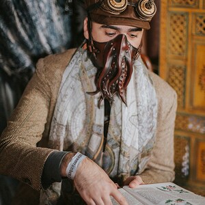 May include: A person wearing a brown leather cap with goggles, a brown leather mask with tentacles, and a white scarf is reading a book with illustrations of plants. The book is open and the person is pointing at the page with their right hand. The person is wearing a brown tweed jacket and a white wristband that says "remarkable".