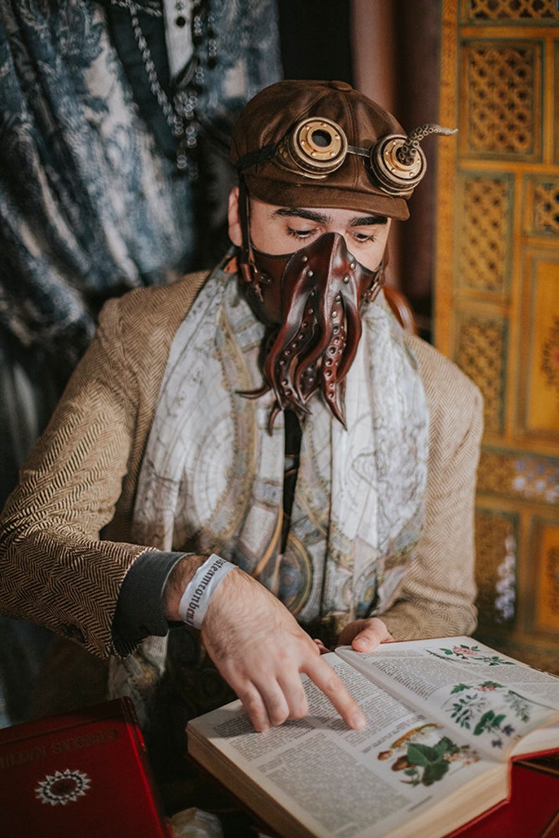 May include: A person wearing a brown leather cap with goggles, a brown leather mask with tentacles, and a white scarf is reading a book with illustrations of plants. The book is open and the person is pointing at the page with their right hand. The person is wearing a brown tweed jacket and a white wristband that says "remarkable".