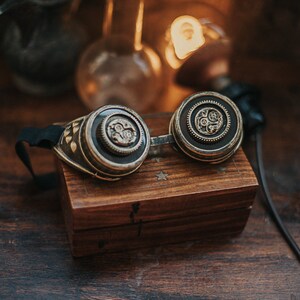 Puede incluir: Un par de gafas steampunk con detalles en lat&oacute;n y negro, que presentan intrincados dise&ntilde;os de engranajes en las lentes. Las gafas est&aacute;n apoyadas en una caja de madera con un dise&ntilde;o de estrella.
