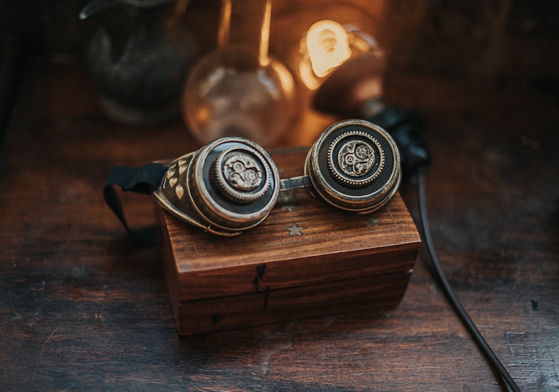 Puede incluir: Un par de gafas steampunk con detalles en lat&oacute;n y negro, que presentan intrincados dise&ntilde;os de engranajes en las lentes. Las gafas est&aacute;n apoyadas en una caja de madera con un dise&ntilde;o de estrella.
