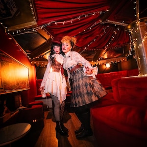 May include: Two people in vintage-inspired costumes pose in a dimly lit room with red velvet seating and draped ceilings. One wears a white dress with a corset, the other a patterned skirt and blouse. String lights add a warm glow.