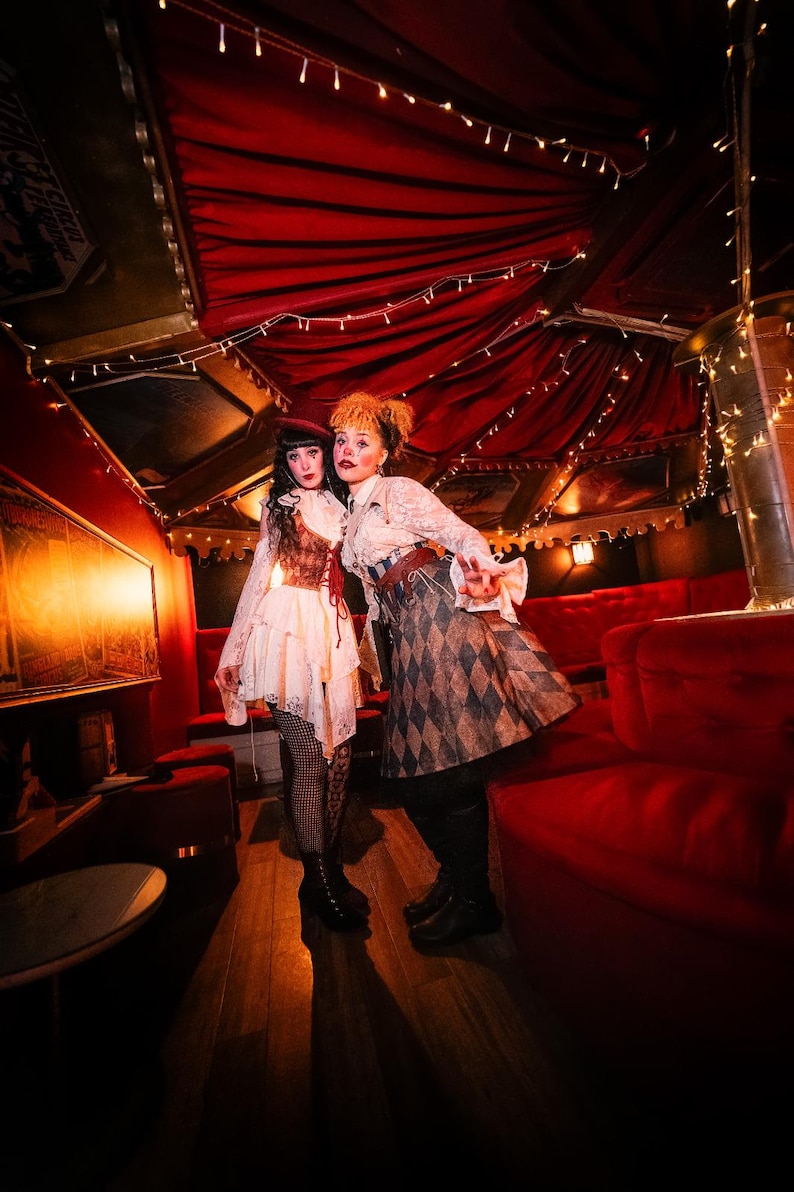 May include: Two people in vintage-inspired costumes pose in a dimly lit room with red velvet seating and draped ceilings. One wears a white dress with a corset, the other a patterned skirt and blouse. String lights add a warm glow.