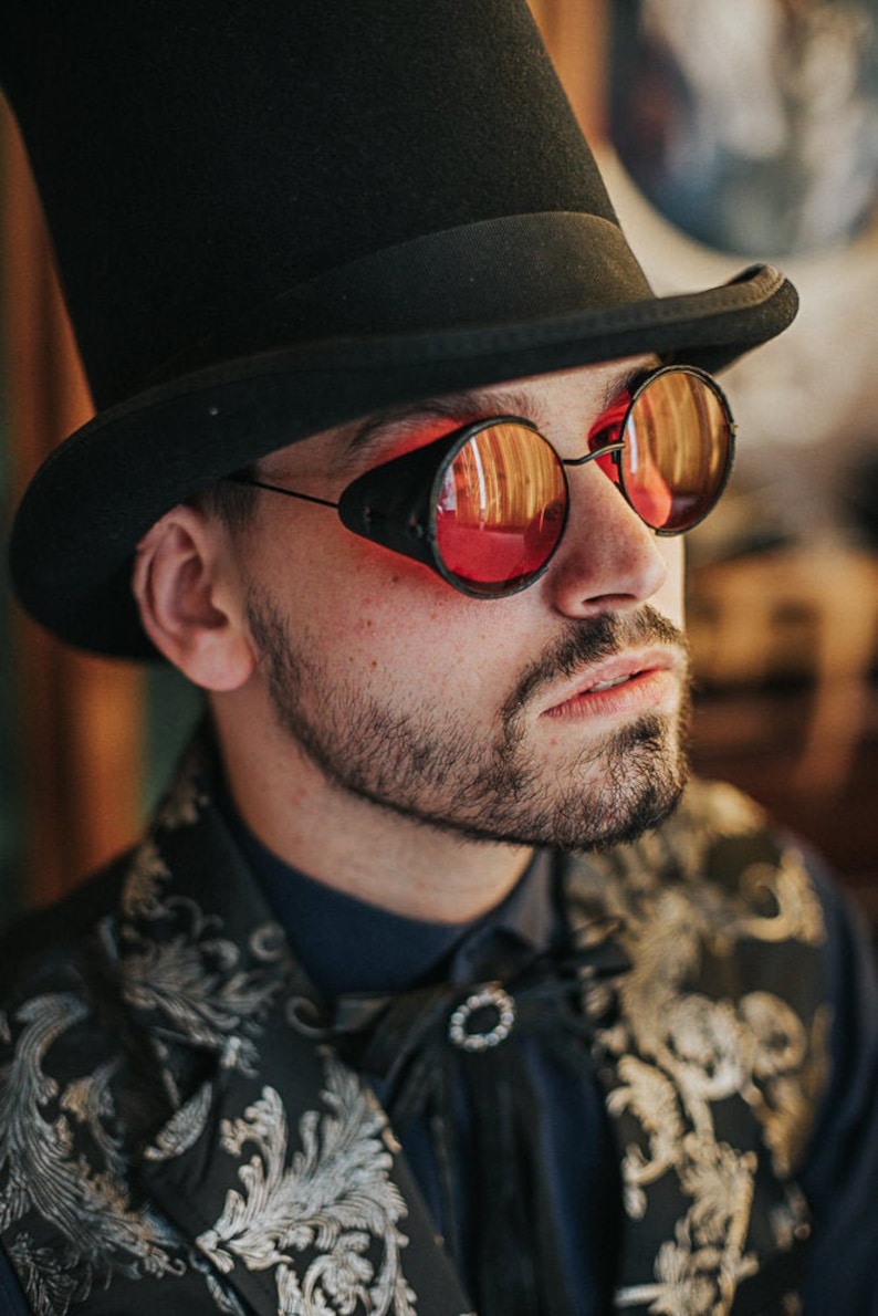 May include: A man wearing a black top hat and round, red-tinted goggles with black frames. He is wearing a black and silver patterned jacket with a black bow tie.