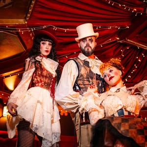 May include: Three people in theatrical costumes stand in a dimly lit space with red curtains and string lights. The costumes include a top hat, beret, ruffled collars, and face makeup. The overall theme is a circus or theatrical performance.