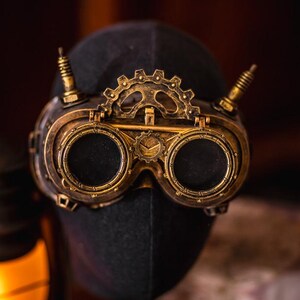 May include: A pair of brass-colored goggles featuring two circular dark lenses, a central gear mechanism, and two vertical spikes on the top frame. The frame has a metallic finish with intricate detailing around the rims and a small gear emblem.