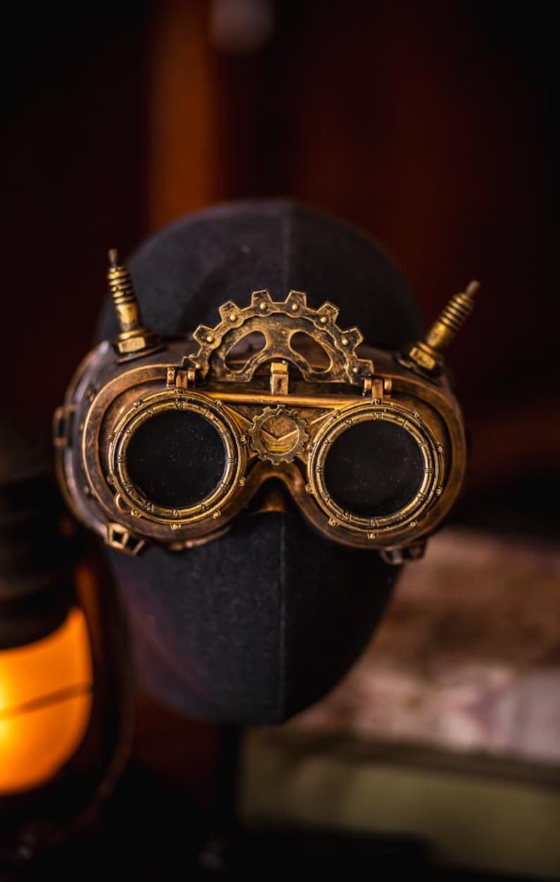 May include: A pair of brass-colored goggles featuring two circular dark lenses, a central gear mechanism, and two vertical spikes on the top frame. The frame has a metallic finish with intricate detailing around the rims and a small gear emblem.