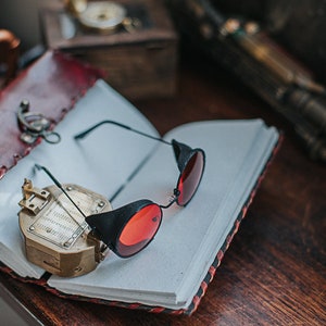 May include: A red leather-bound journal with a compass and a pair of black steampunk-style glasses with red lenses.
