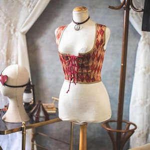 May include: A corset-style top on a mannequin, featuring a red and tan diamond pattern. The top has a lace-up front and a burgundy choker necklace. A head form with a heart-shaped eye mask is visible in the background. The scene evokes a vintage aesthetic.