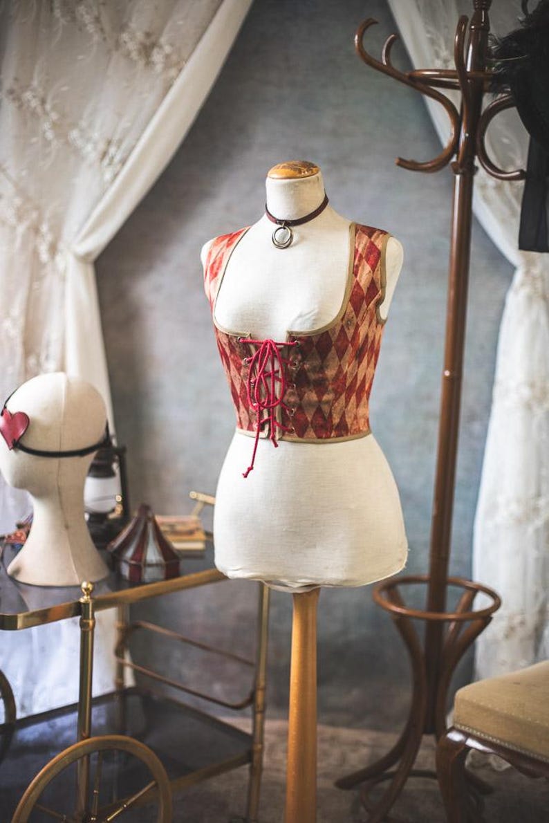 May include: A corset-style top on a mannequin, featuring a red and tan diamond pattern. The top has a lace-up front and a burgundy choker necklace. A head form with a heart-shaped eye mask is visible in the background. The scene evokes a vintage aesthetic.
