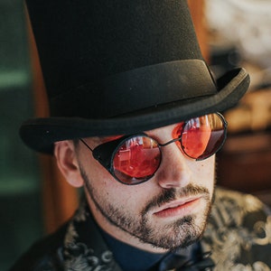 May include: A man wearing a black top hat and red-tinted round glasses with a black frame. He is wearing a black and gold patterned vest over a blue shirt.