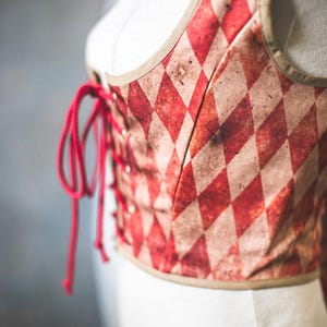 May include: A red and cream-colored corset with a diamond pattern. The corset has a lace-up front with red lacing and a tan trim. The garment is displayed on a white mannequin.