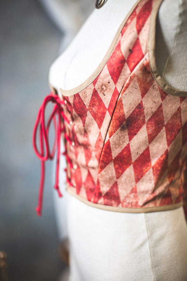 May include: A red and cream-colored corset with a diamond pattern. The corset has a lace-up front with red lacing and a tan trim. The garment is displayed on a white mannequin.