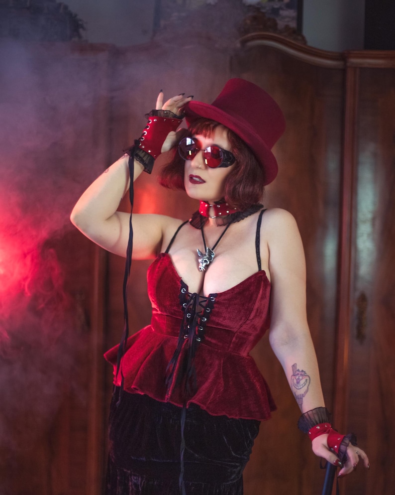 May include: A woman wearing a red velvet corset with black lace-up detailing, a black skirt, and a red top hat. She is also wearing red leather gloves and red sunglasses. The background is a dark, smoky room.