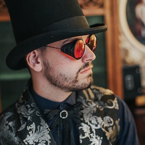 May include: A man wearing a black top hat, a black and silver patterned vest, a dark blue shirt, and red-tinted goggles.
