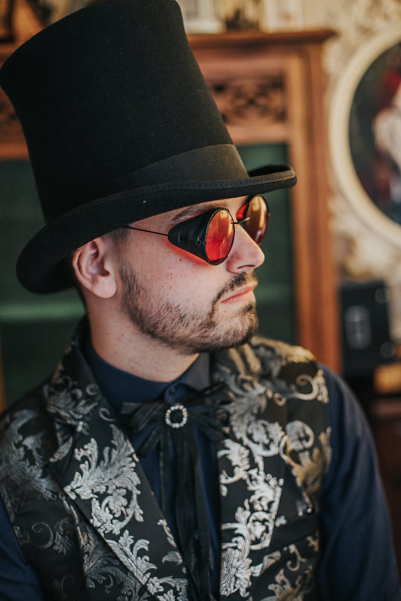May include: A man wearing a black top hat, a black and silver patterned vest, a dark blue shirt, and red-tinted goggles.