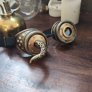 May include: A pair of steampunk goggles with a brass finish and a black strap. One lens has a tentacle design.