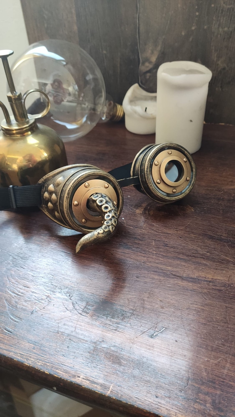May include: A pair of steampunk goggles with a brass finish and a black strap. One lens has a tentacle design.