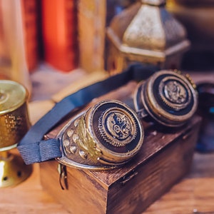 Puede incluir: Un par de gafas steampunk con detalles en lat&oacute;n y negro. Las gafas tienen un dise&ntilde;o circular con engranajes y otros detalles intrincados. Las gafas est&aacute;n sobre una caja de madera.