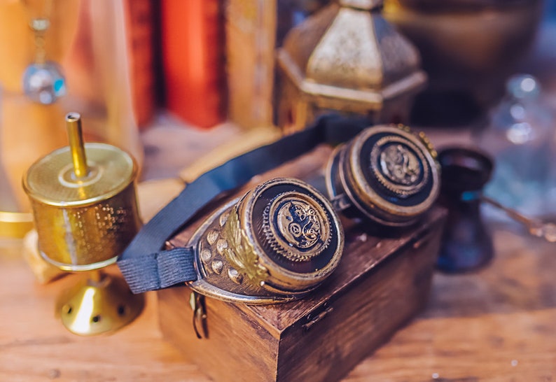 Puede incluir: Un par de gafas steampunk con detalles en lat&oacute;n y negro. Las gafas tienen un dise&ntilde;o circular con engranajes y otros detalles intrincados. Las gafas est&aacute;n sobre una caja de madera.