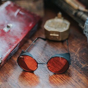 May include: Round, red-tinted sunglasses with black frames. The sunglasses are lying on a wooden surface.