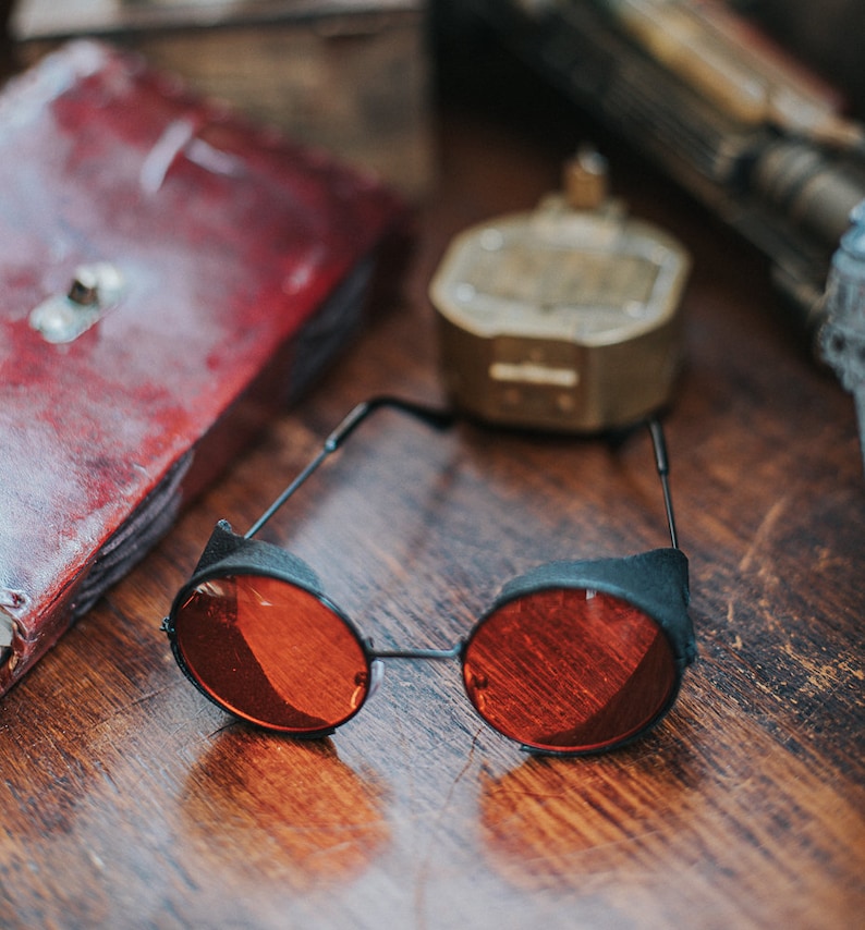 May include: Round, red-tinted sunglasses with black frames. The sunglasses are lying on a wooden surface.