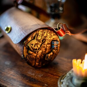 May include: A cylindrical leather bag with a gold embossed design featuring an Egyptian hieroglyphic pattern and a small golden figure. The bag has a brown leather strap with a brass buckle.