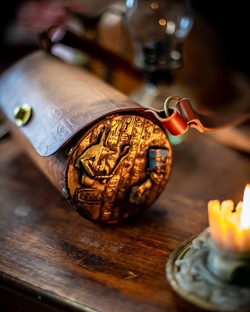 May include: A cylindrical leather bag with a gold embossed design featuring an Egyptian hieroglyphic pattern and a small golden figure. The bag has a brown leather strap with a brass buckle.