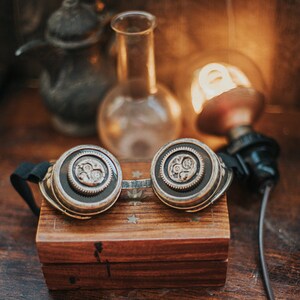 Puede incluir: Un par de gafas steampunk con detalles en lat&oacute;n y plata, descansando sobre una caja de madera con un patr&oacute;n de estrellas. Las gafas tienen una correa de cuero negro y est&aacute;n enfocadas, mientras que el fondo est&aacute; desenfocado.